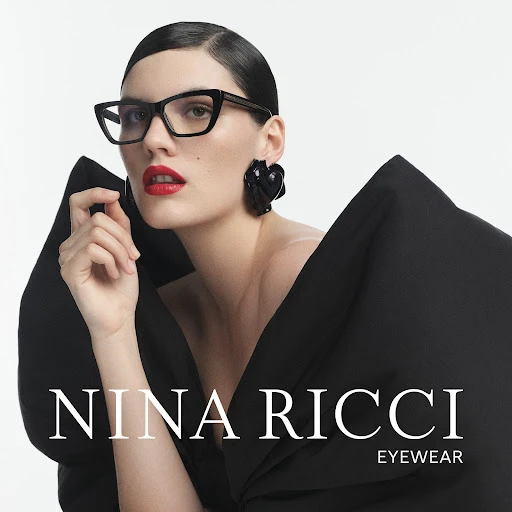 A woman with dark hair and red lipstick models black eyeglasses, wearing a black dress and black earrings against a white backdrop.