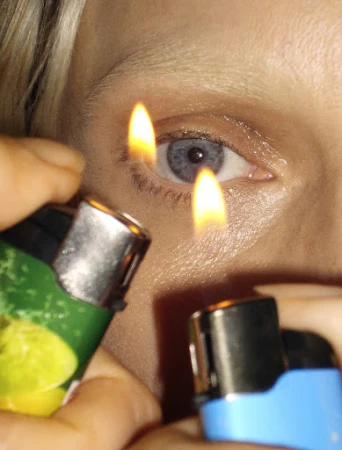 Close-up portrait of a blue eye with two lit lighters held near the face by manicured hands.