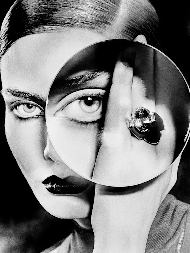 Monochrome beauty shot featuring a woman with dark lipstick looking through a circular reflector, highlighting an eye and a finger ring.
