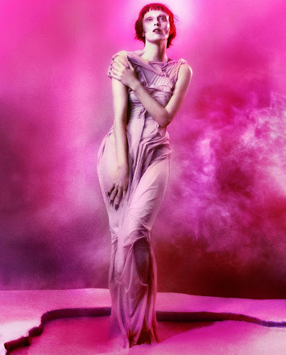 Elegant woman in sheer dress poses against a pink backdrop, illuminated by a soft glow, emphasizing her sophisticated beauty.