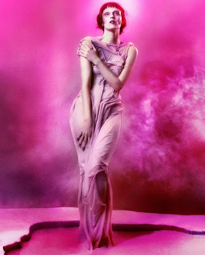 Elegant woman in sheer dress poses against a pink backdrop, illuminated by a soft glow, emphasizing her sophisticated beauty.