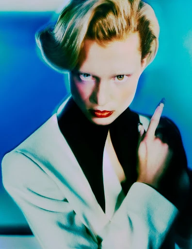 Woman with platinum blonde hair, wearing a white blazer and red lipstick, gazes at the camera while gesturing with her manicured finger.