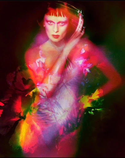 Surreal portrait of a red-haired woman in a white dress amidst colorful abstract floral elements with a dark studio backdrop and soft focus.