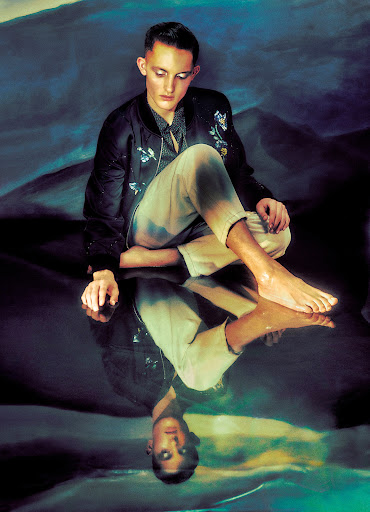 A young adult sits cross-legged on a reflective surface in front of a painted backdrop, appearing pensive with a reflected image.