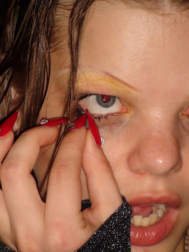 Close-up of a young woman with red nails holding her eyelid open, with visible make-up and a dark top, shot indoors.