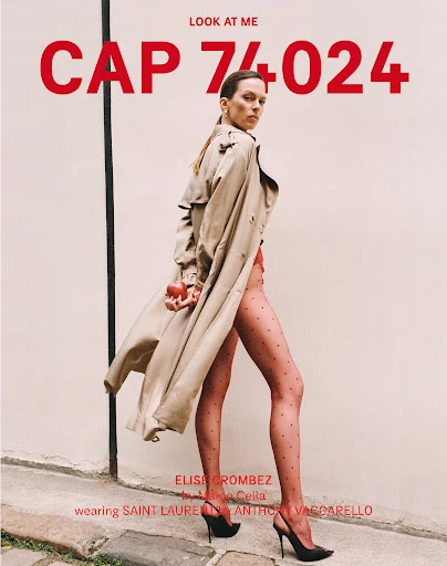 Fashion editorial shot of a female model wearing a beige trench coat and high heels, holding a red apple in front of a beige wall.