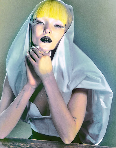 Dramatic studio portrait of a yellow-haired female wearing white shawl and expressive makeup, posing with folded hands at a wooden table.