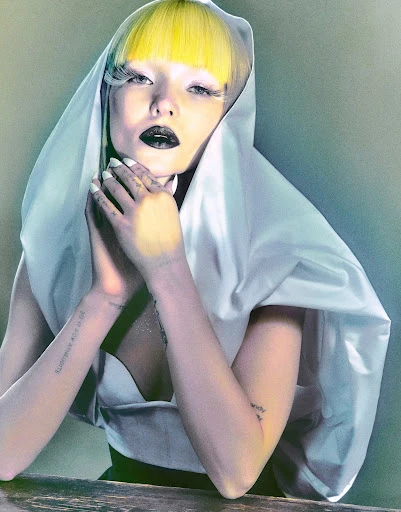 Dramatic studio portrait of a yellow-haired female wearing white shawl and expressive makeup, posing with folded hands at a wooden table.