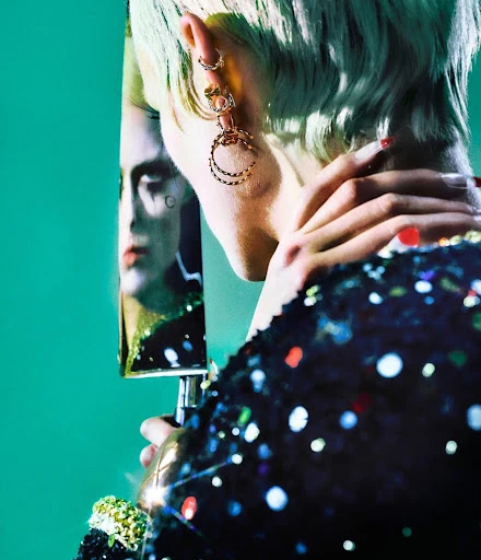 Stylish woman with a shimmering black dress and short teal hair poses, wearing hoop earrings, with a partial reflection on a teal background.