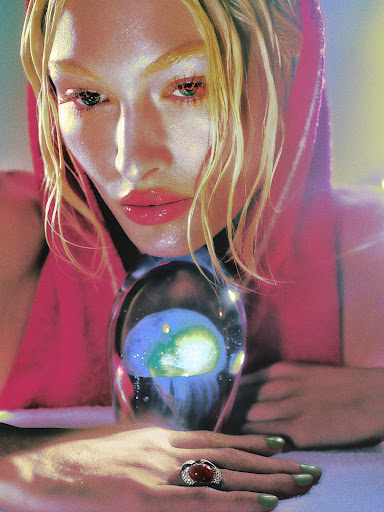 Glamorous woman with blonde hair in pink hooded top holding glass orb with planetary image, wearing large jeweled ring, dramatic lighting.