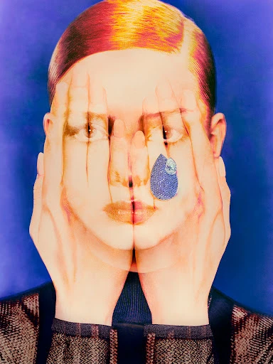 Conceptual portrait of a person obscuring their face with hands, featuring a blue gemstone teardrop and colorful hair against a blue backdrop.