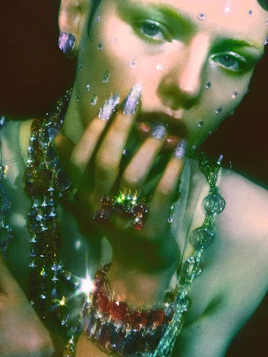 Close-up studio shot of a woman with sparkling gems on her face, heavy beaded jewelry, and lavender nails posing with hand near her face.