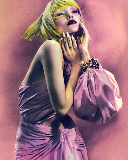 A woman with fair skin and bright yellow hair poses in a pink gown, set against a muted pink background in a fashion shot.