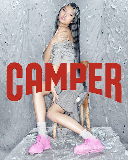 Asian woman poses in stylish pink shoes and an ensemble of gray separates, seated on a vintage chair against a textured backdrop.