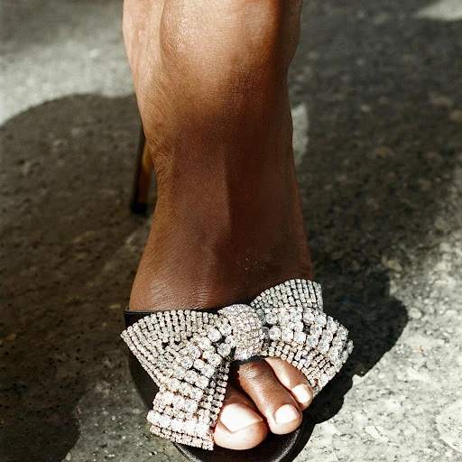 Close-up of woman's foot in a black sandal with a large rhinestone bow, set against a concrete backdrop with shadows.