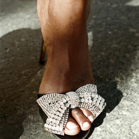 Close-up of woman's foot in a black sandal with a large rhinestone bow, set against a concrete backdrop with shadows.