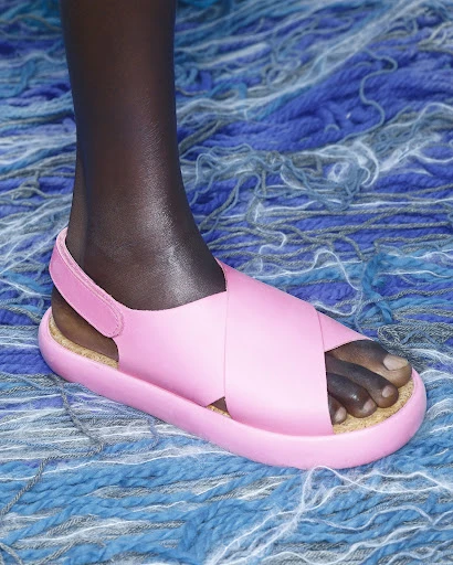 Close-up of a pink sandal on the foot of a dark-skinned person, set against a background of blue and gray yarn strands.