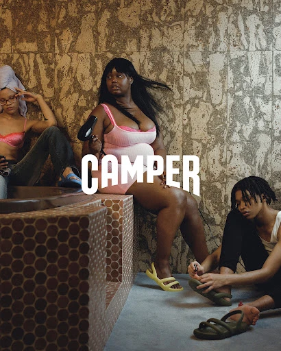 Three young adults in casual fashion sit in a tiled spa-like setting; one woman uses a hairdryer; all wear 'Camper' branded slides.