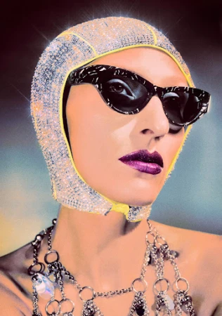 Close-up portrait of a woman wearing a sparkling rhinestone hood, cat-eye sunglasses, and metallic chain body jewelry.