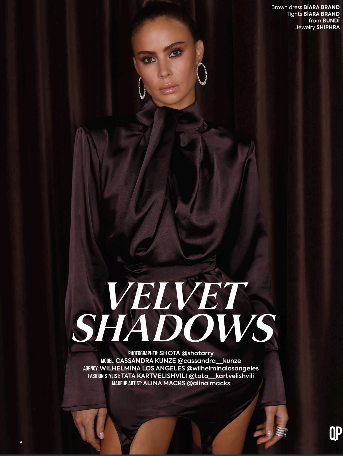 A woman poses in a brown satin dress with a dramatic bow, complemented by hoop earrings, against a dark, velvety backdrop for a fashion editorial.