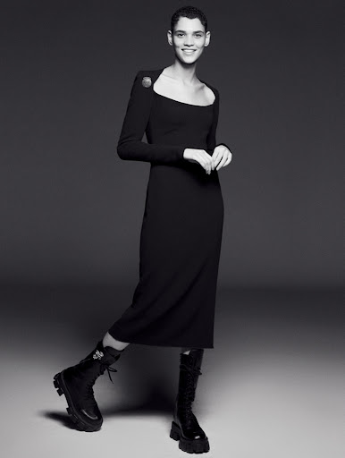 A woman wearing a long-sleeved black midi dress and black combat boots poses in a studio with a neutral grey background.