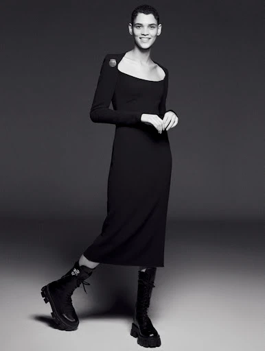 A woman wearing a long-sleeved black midi dress and black combat boots poses in a studio with a neutral grey background.