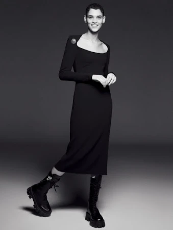 A woman wearing a long-sleeved black midi dress and black combat boots poses in a studio with a neutral grey background.