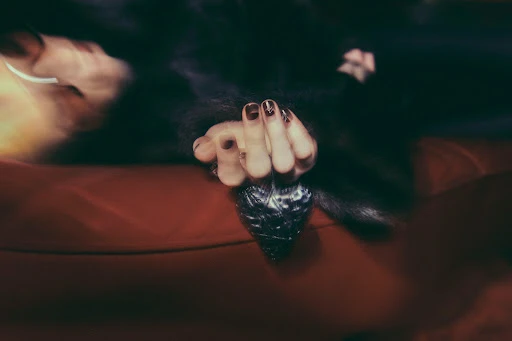 A hand with decorated nails holds a textured, dark heart object, set against a blurred backdrop of dark hair and a red surface.