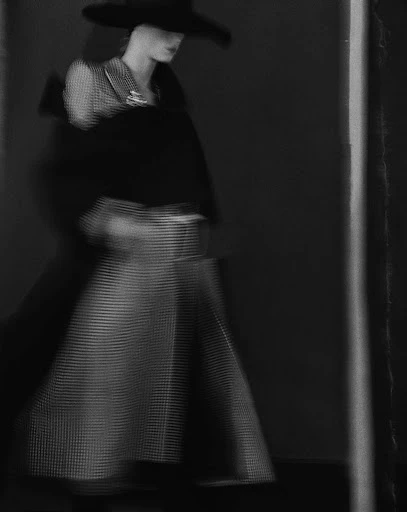 An adult female in a black hat and patterned skirt poses in a high-fashion shot with motion blur, indoors against a dark background.