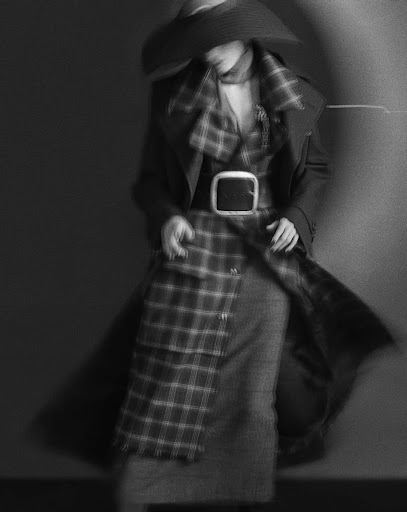 Black and white fashion portrait of a woman in a plaid dress, coat, hat, and belt in a blurred motion with a spotlight effect.