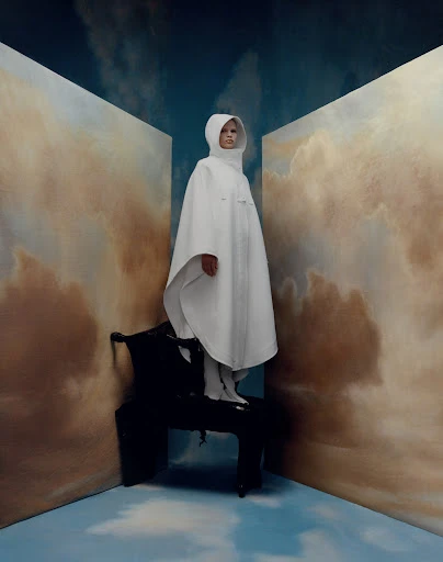 Model in white hooded poncho on a black chair against a cloudy backdrop in a surreal fashion editorial shot. Fashion photography.