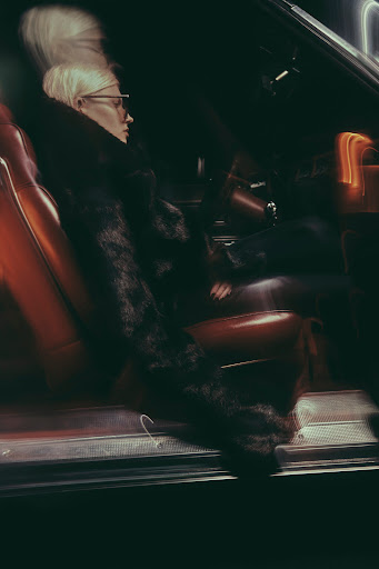 Blonde woman with glasses seated in the dark leather interior of a car at night with motion blur lights.