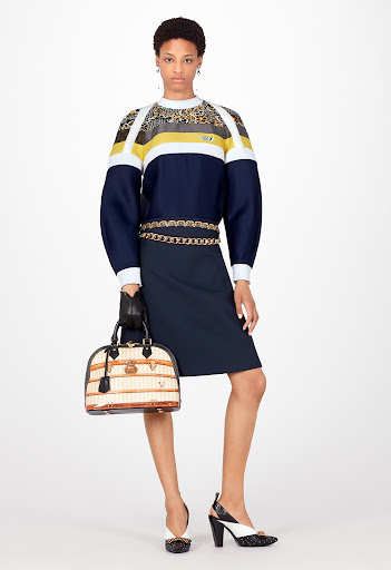 Fashion shot of a Black female model in a color-block top and navy skirt, accessorized with a chain belt and handbag, against a white backdrop.