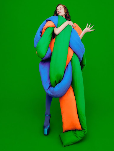 Fashionable woman in padded blue, green, and orange outerwear and matching tights poses against a vibrant green background.