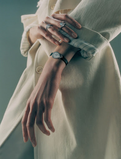 Close-up of an adult female's hands with rings and a watch on a wrist, wearing a white coat against a soft green backdrop.