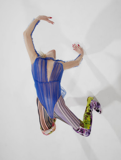 Overhead shot of a woman in a blue bodysuit and floral patterned leggings, arms raised against a stark white background.