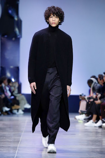 Runway fashion show featuring a curly-haired adult male model wearing a long black coat and dark pants, walking toward the camera.