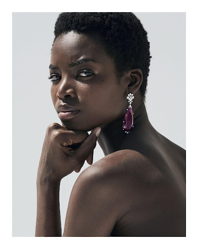 Close-up portrait of a Black woman with short dark hair wearing a large teardrop gemstone earring against a light grey background.