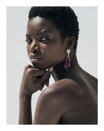 Close-up portrait of a Black woman with short dark hair wearing a large teardrop gemstone earring against a light grey background.