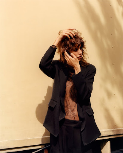 Model with tousled brown hair wearing a black blazer and sheer patterned top posing outdoors