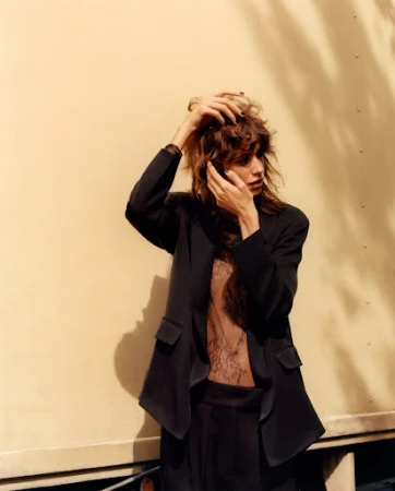 Model with tousled brown hair wearing a black blazer and sheer patterned top posing outdoors
