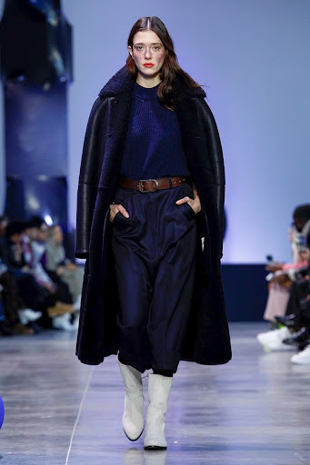 Model walking the runway wearing a long black coat over a blue sweater and dark trousers with light boots