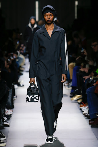 Runway fashion show featuring a Black female model in a dark jumpsuit with white stripes, holding a black and white Y-3 handbag.