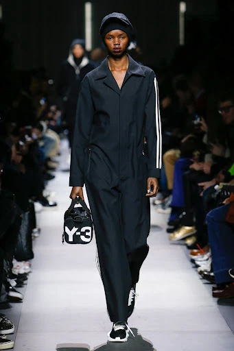 Runway fashion show featuring a Black female model in a dark jumpsuit with white stripes, holding a black and white Y-3 handbag.