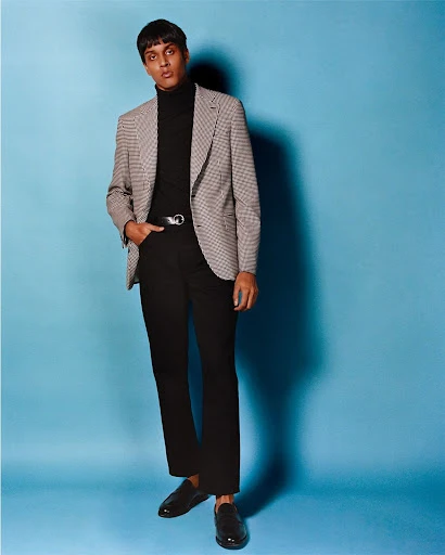 Full-length shot of a young adult man wearing a grey houndstooth blazer, black turtleneck and pants, and black dress shoes on blue backdrop.