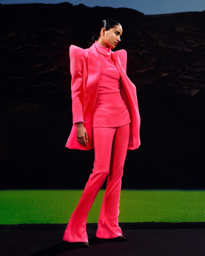 Woman in a hot pink suit poses in front of a dark mountain landscape with a neon green strip highlighting the ground plane.