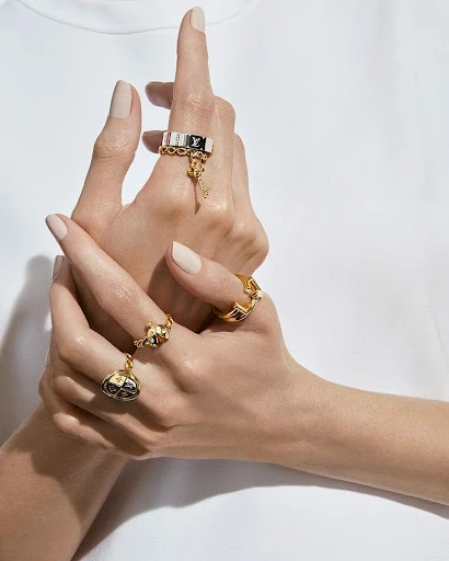 Close-up of adult female hands with rings, wearing gold and black Louis Vuitton rings on a white surface in bright, soft lighting.