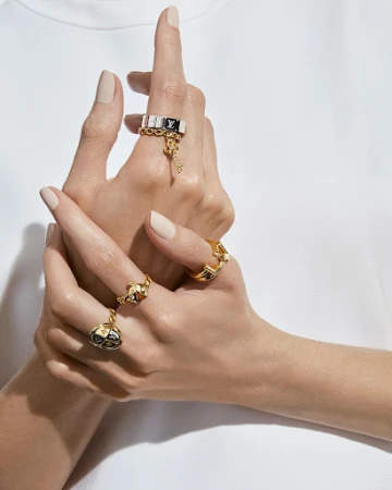 Close-up of adult female hands with rings, wearing gold and black Louis Vuitton rings on a white surface in bright, soft lighting.