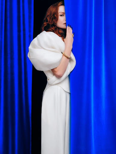 Elegant woman in flowing white gown stands near a royal-blue curtain, hands in a poised position and looking off camera.