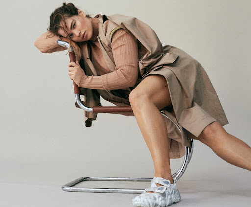 A brunette woman in a beige coat and textured top, seated on a silver metal chair in a relaxed pose, offset against a neutral backdrop.